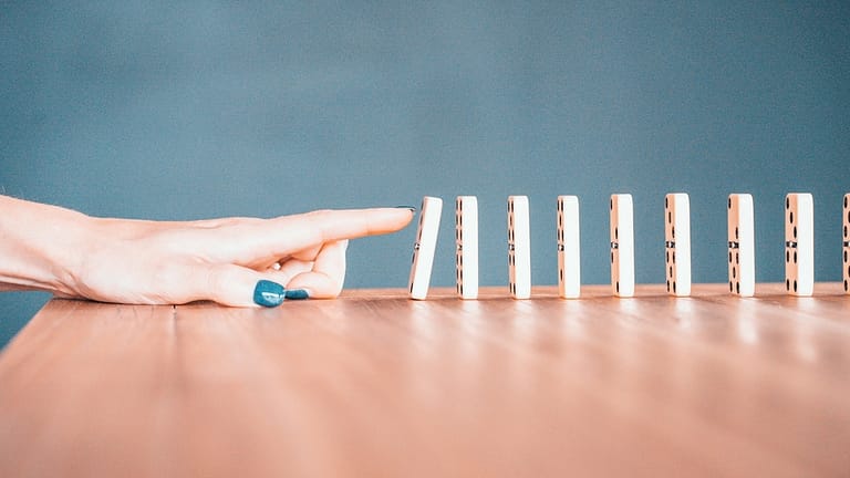 Image: A woman's hand, nails painted blue, knocking down the first domino in a line of many. Symbolic for taking the first step to achieving her goals.