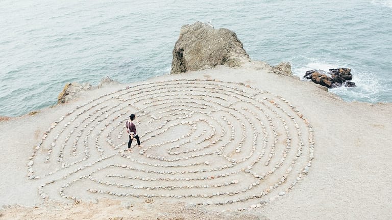 Image: A woman walking through a circular rock maze on a beach near the ocean, taking time for herself to tackle mental load.