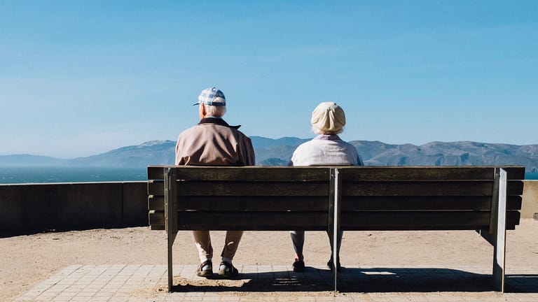 Image: Two elderly individuals, a couple, sitting on a bench.