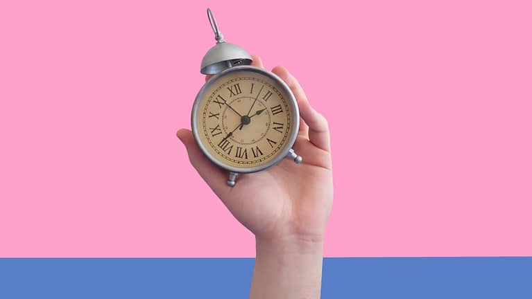 Image: A hand holding an analog clock in front of a pink and blue background, a metaphor for time blocking.