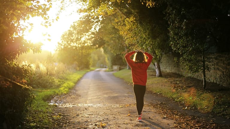 Image: A woman relieves stress by walking along a path with her hands on her head, the sun shining.