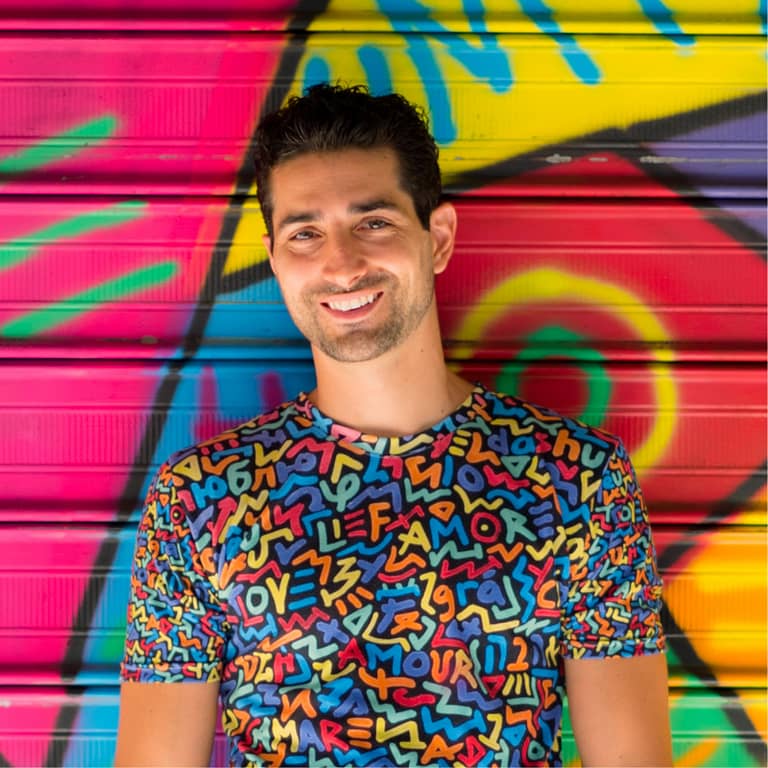 Artist Marco Santini stands smiling in front of one of his vibrantly colored 'One Love' murals, wearing a shirt with the same theming: the word love is spelled out in multiple colors against a black background