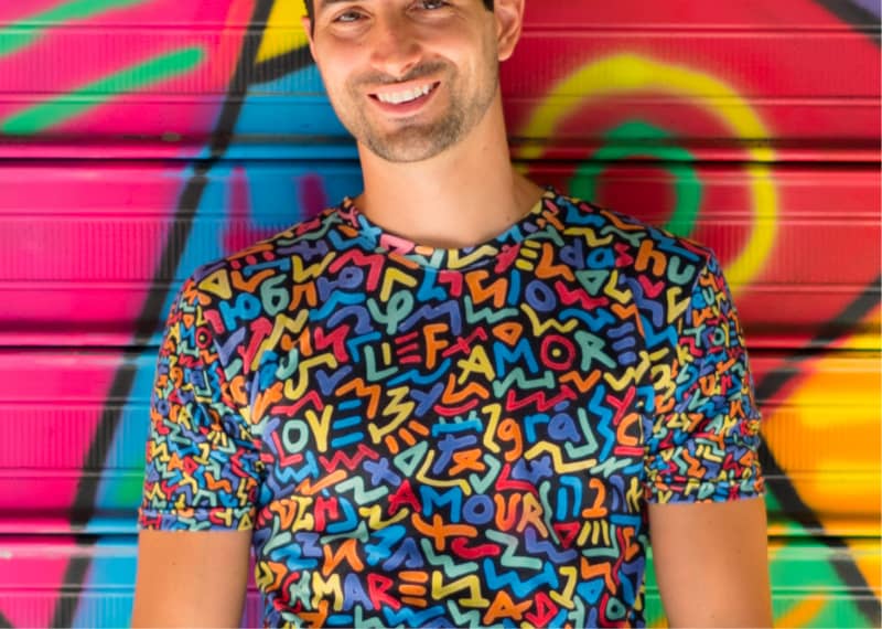 Artist Marco Santini stands smiling in front of one of his vibrantly colored 'One Love' murals, wearing a shirt with the same theming: the word love is spelled out in multiple colors against a black background