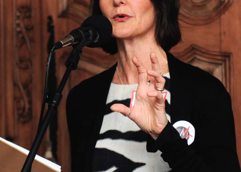 Liz Stookey Sunde, co founder of music to life, speaks into a large microphone. She is gesturing a grasp with one hand and holding a book in the other.