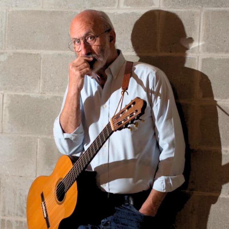 Noel 'Paul' Stookey of the iconic musical trio 'Peter, Paul, and Mary' stands against a cinderblock wall with a guitar.