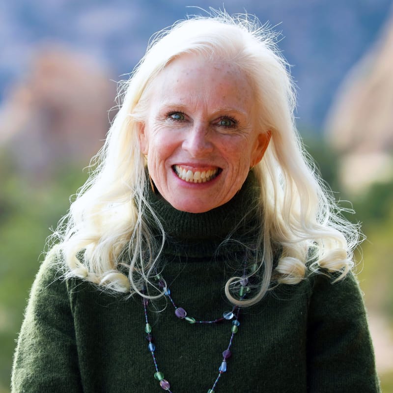 Author, Relationship Coach, and Podcaster Rebecca Mullen, smiles against a background of blurred outdoor scenery. She is a white woman with platinum-blonde hair, wearing a dark green turtleneck and a long necklace of blue beads.