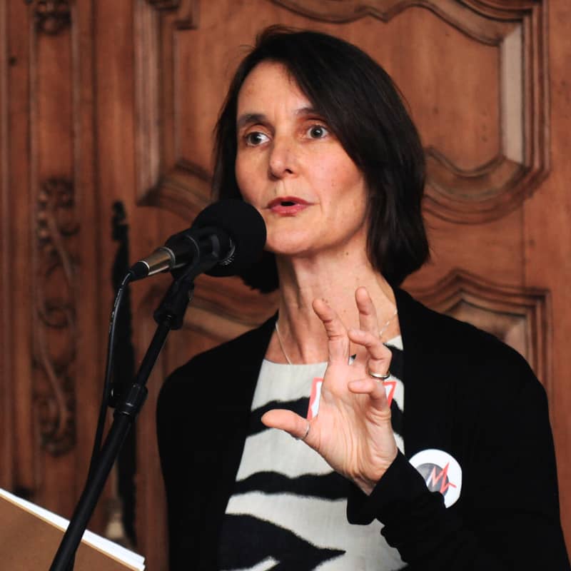 Liz Stookey Sunde, co founder of music to life, speaks into a large microphone. She is gesturing a grasp with one hand and holding a book in the other.