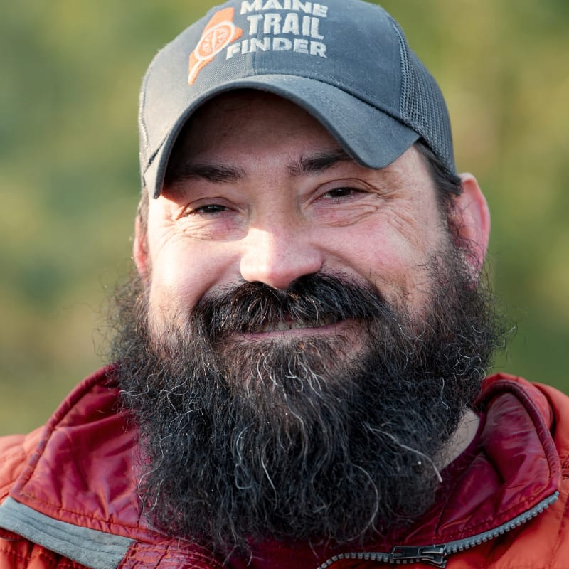 Enock Glidden, adventurer, outdoorsman, and disabled athlete, smiles at the camera against a blurred leafy backdrop.