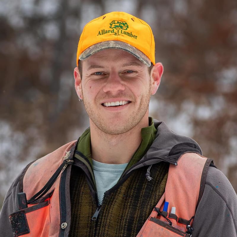 Ethan Tapper, Forester and Author, stands in a coat and orange vest wearing a yellow ball cap and smiling. He stands against a blurred, wooded background.