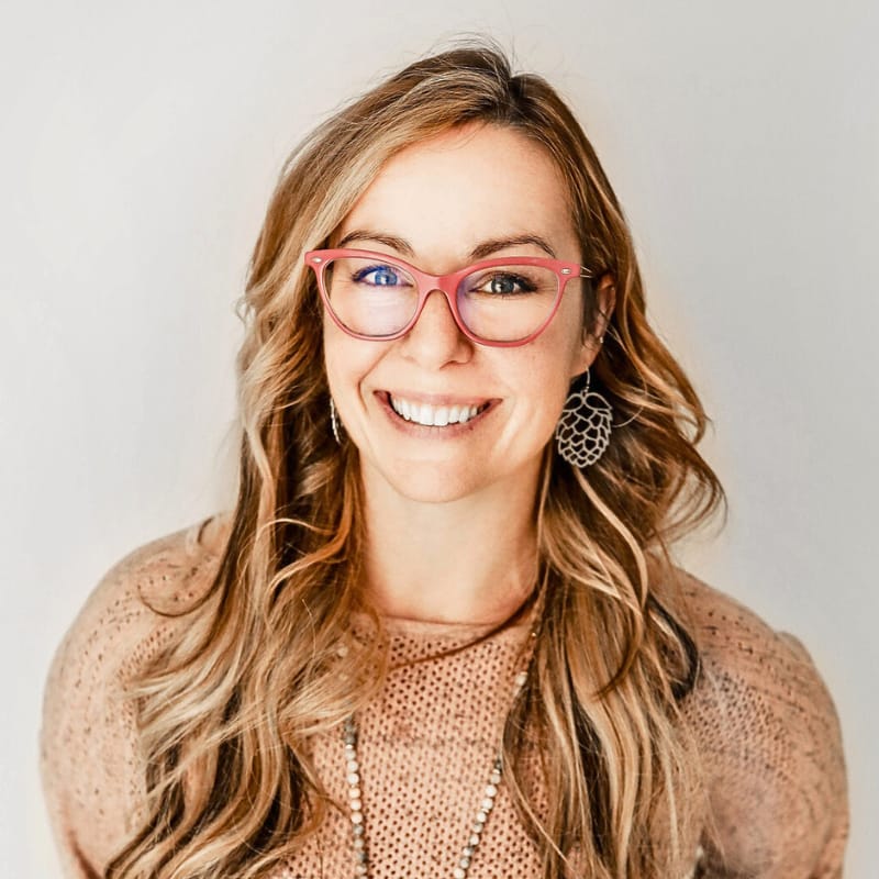 a picture of corrin thompson, marketing specialist for thompson rv. She is a white woman with long, wavy blond hair wearing reddish-pink, rectangular glasses, a tan sweater, and a long silver necklace. She smiles against a white gray gradient background