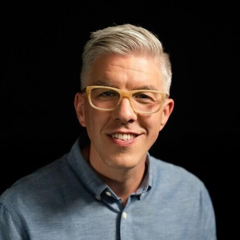 Mark Champagne, Author, Consultant, and Podcast Host, smiles against a black background. He is a white man with warm-toned skin and short, styled gray hair, wearing yellow eyeglasses and a denim-colored button-down shirt