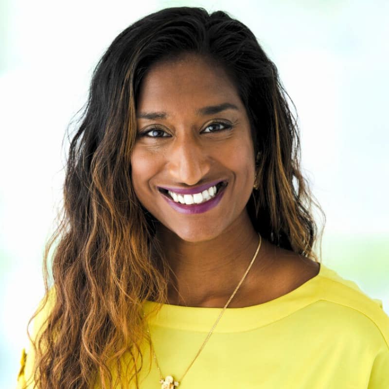 Sabrina Phillips, a woman with warm-toned brown skin and black-to-reddish-blonde balayage hair, smiles at the viewer directly. She wears a wide-collared yellow shirt and is standing against a very bright background with hints of light green.