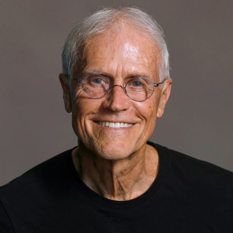 Paul Hawken smiles at the viewer directly,. He is an older man with warm-toned, light-colored skin and short white hair, wearing ovular wire-rimmed glasses and a black shirt, contrasting with a warm gray background. Paul Hawken joins The Goodness Exchange podcast today to discuss how carbon is not the climate change enemy we think it is.