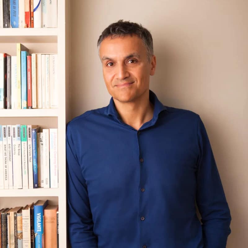 Carl Honore leans casually against a white bookshelf and smiles at the viewer. He has warm-toned skin and short black hair, shorter and grayer on the sides. He wears a navy blue button-down shirt.