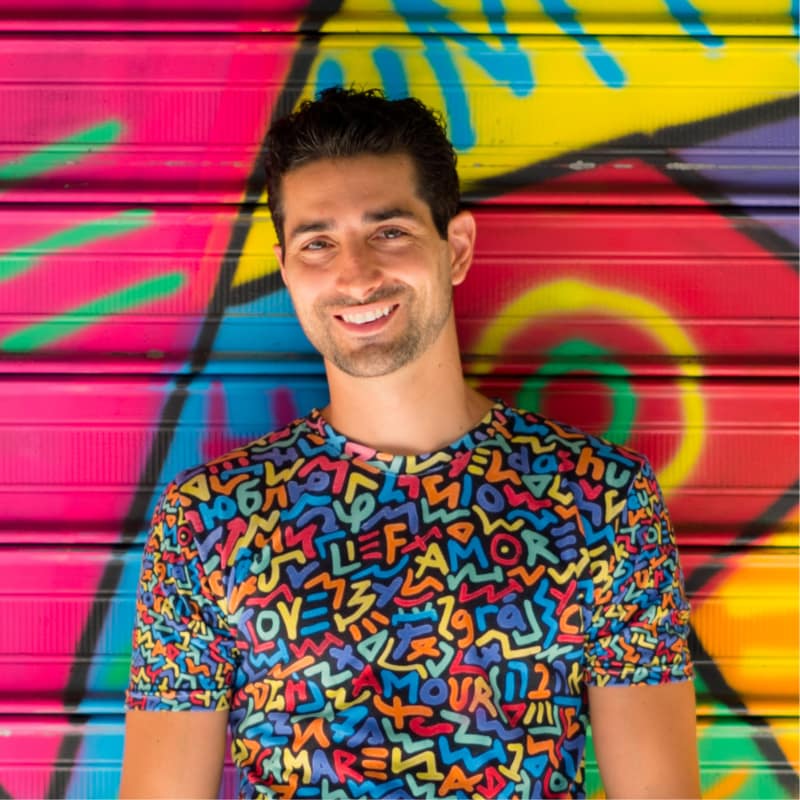 Artist Marco Santini stands smiling in front of one of his vibrantly colored 'One Love' murals, wearing a shirt with the same theming: the word love is spelled out in multiple colors against a black background