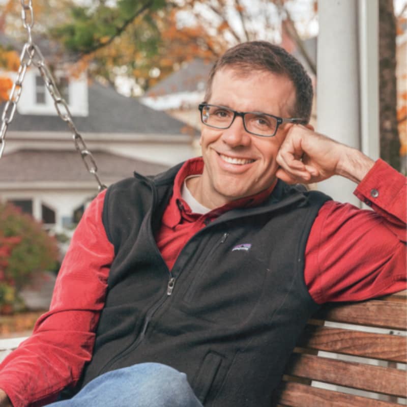 Michael Wood-Lewis, founder of front porch forum, sitting on a front porch swing against a suburban autumn backdrop.