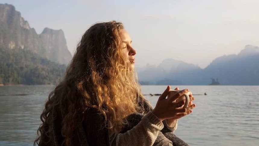 Image: A woman holding a cup of tea with both hands. In the background, a beautiful lake backed by mountains. She is looking towards the sun and smiling. How to control your emotions in any situation.