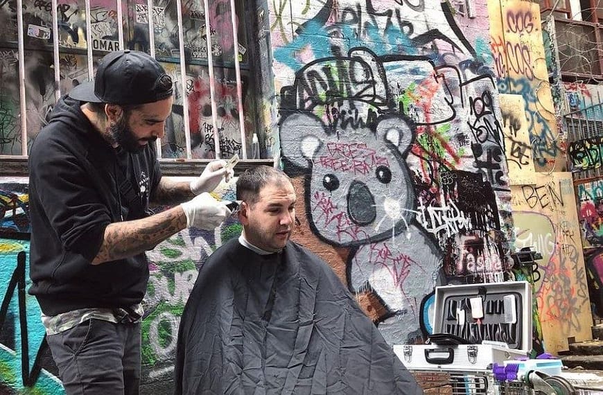 Image: The Streets Barber giving a haircut outside, next to a wall covered in beautiful graffiti