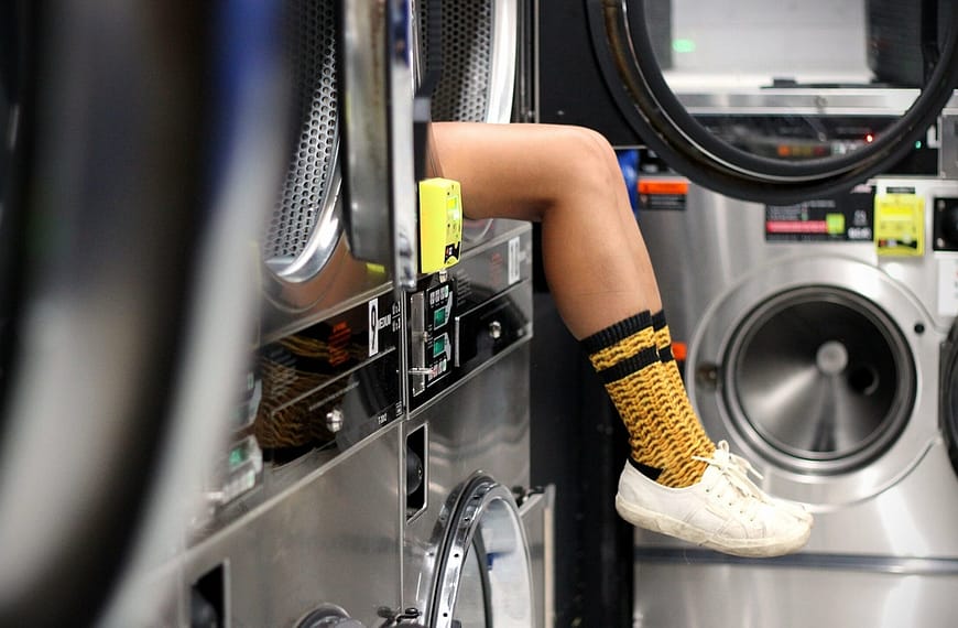 A pair of legs sticking out of a dryer, wearing long yellow socks and white shoes, at a laundromat.