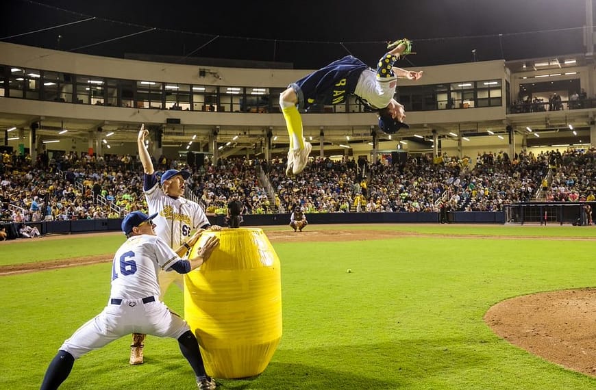 Image: A player for the Savannah Bananas doing a backflip off of a barrel in front of a cheering crowd.