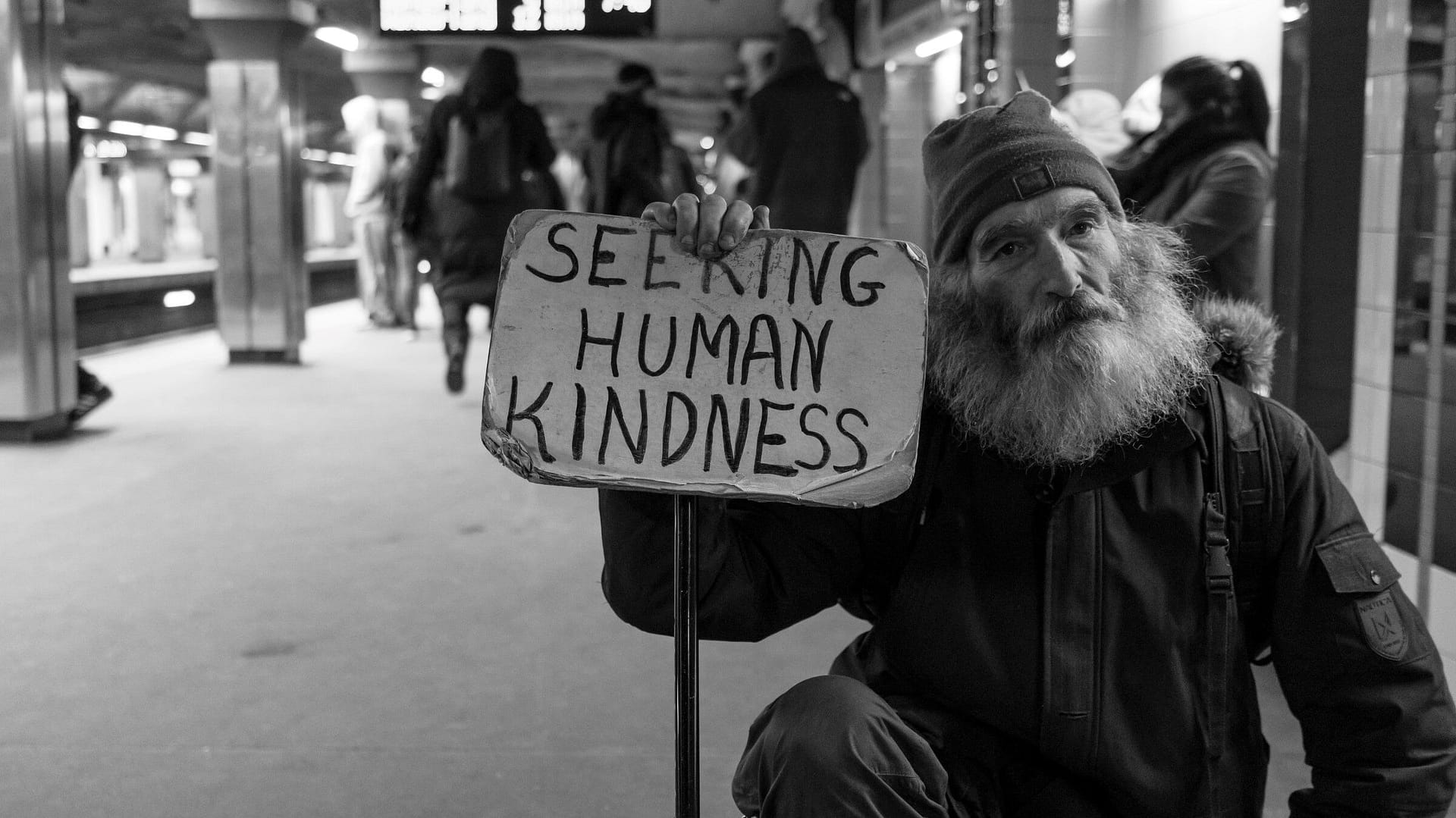 Image: Man holding a sign that says "seeking human kindness" in a Boston subway platform