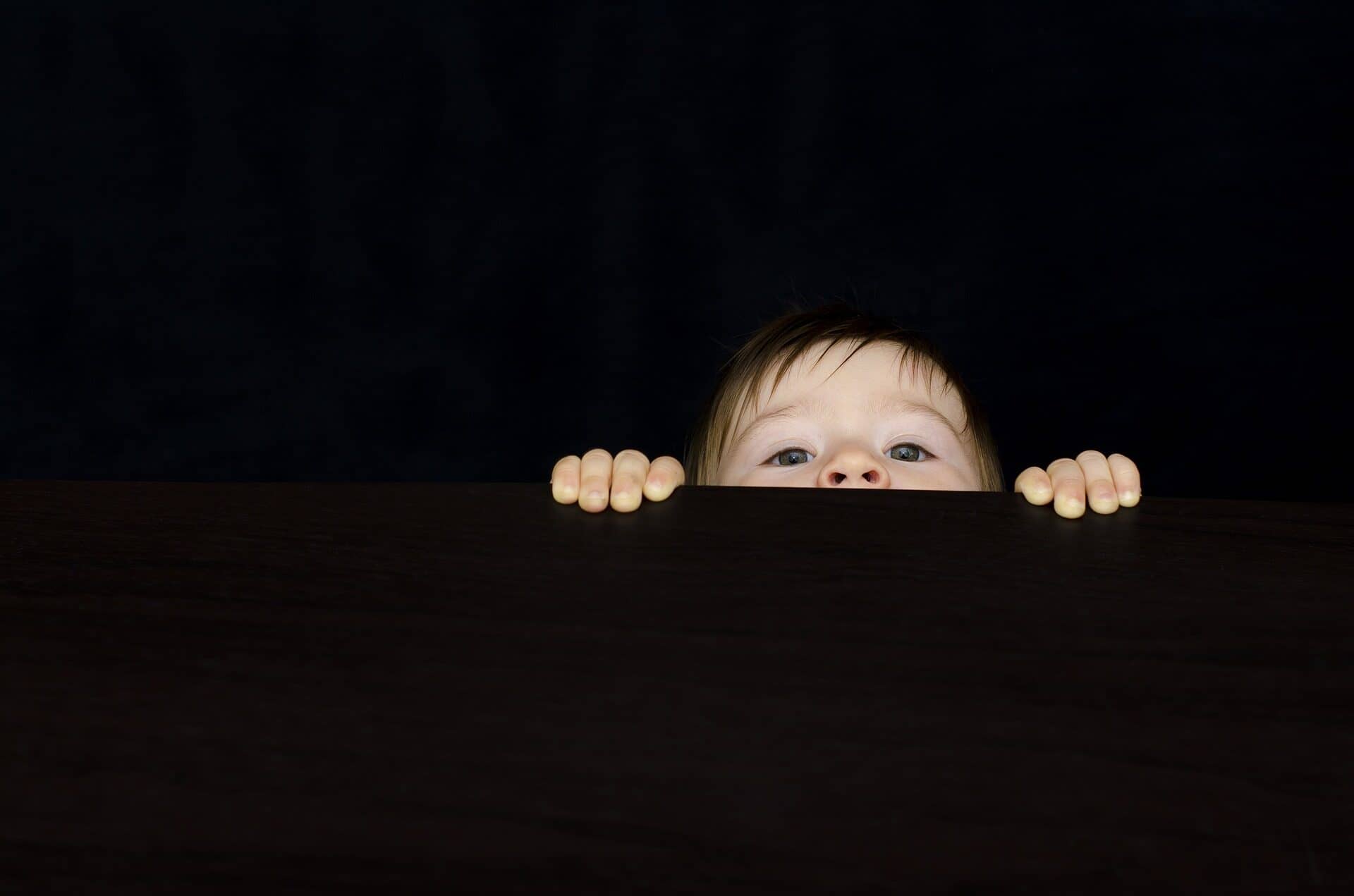 Image: A little boy peeking over a black wall, only eyes, nose and hands showing.