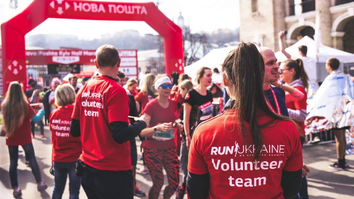 Image: A woman standing among volunteers at a charity foot race, facing away from the camera wearing a red shirt that says "volunteer team".