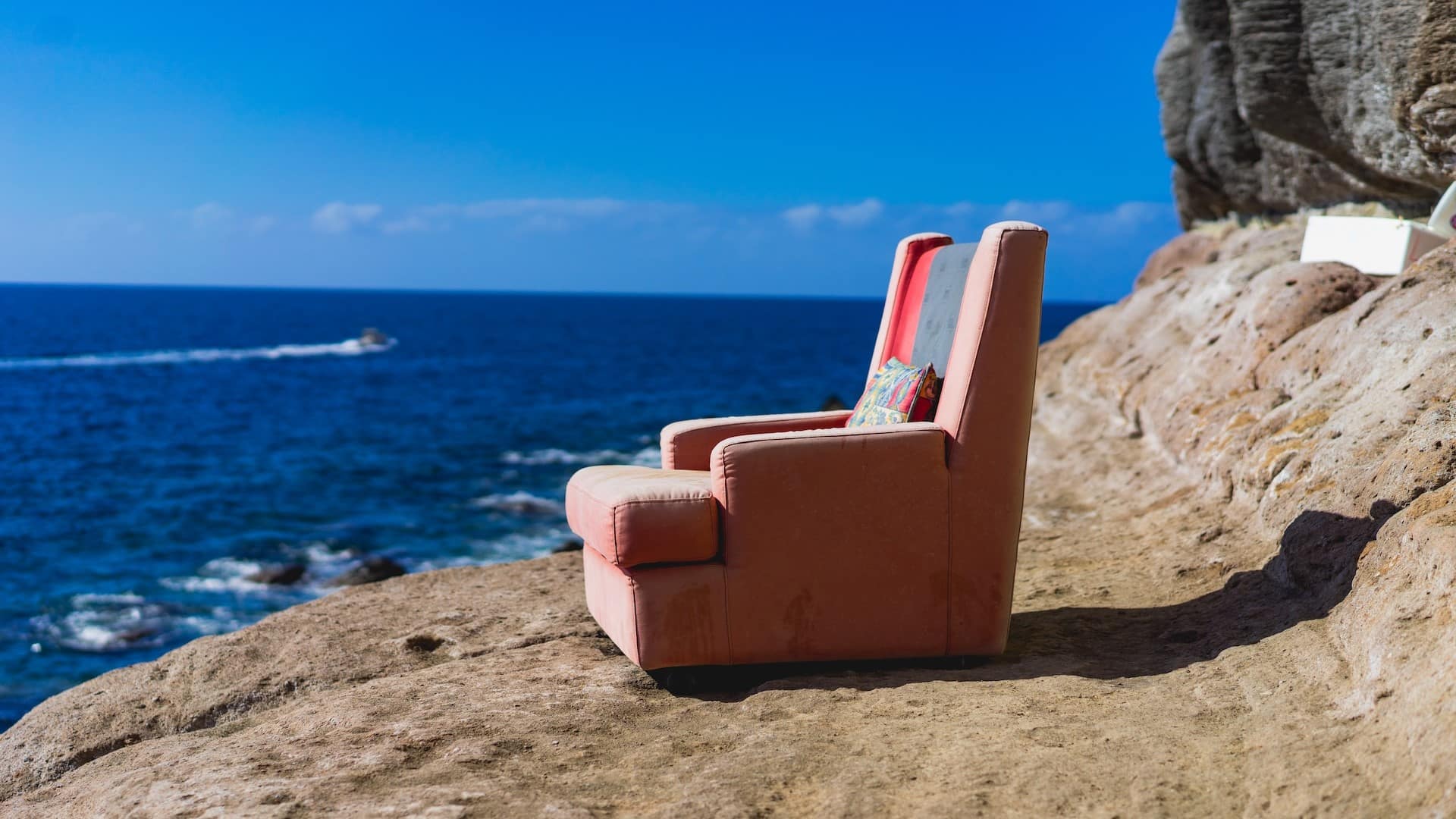 Image: A pink comfy chair, out of place, sitting on a rock by a beach. It asks the question: what else could we do from the couch, other than watch the news?
