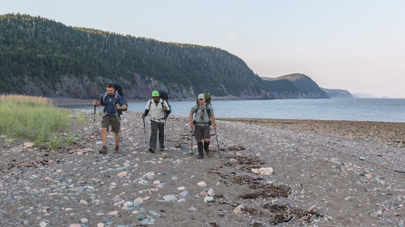 Image: Three men walk across a stone covered beach in on the Fundy Footpath with the Bay of Fundy behind them