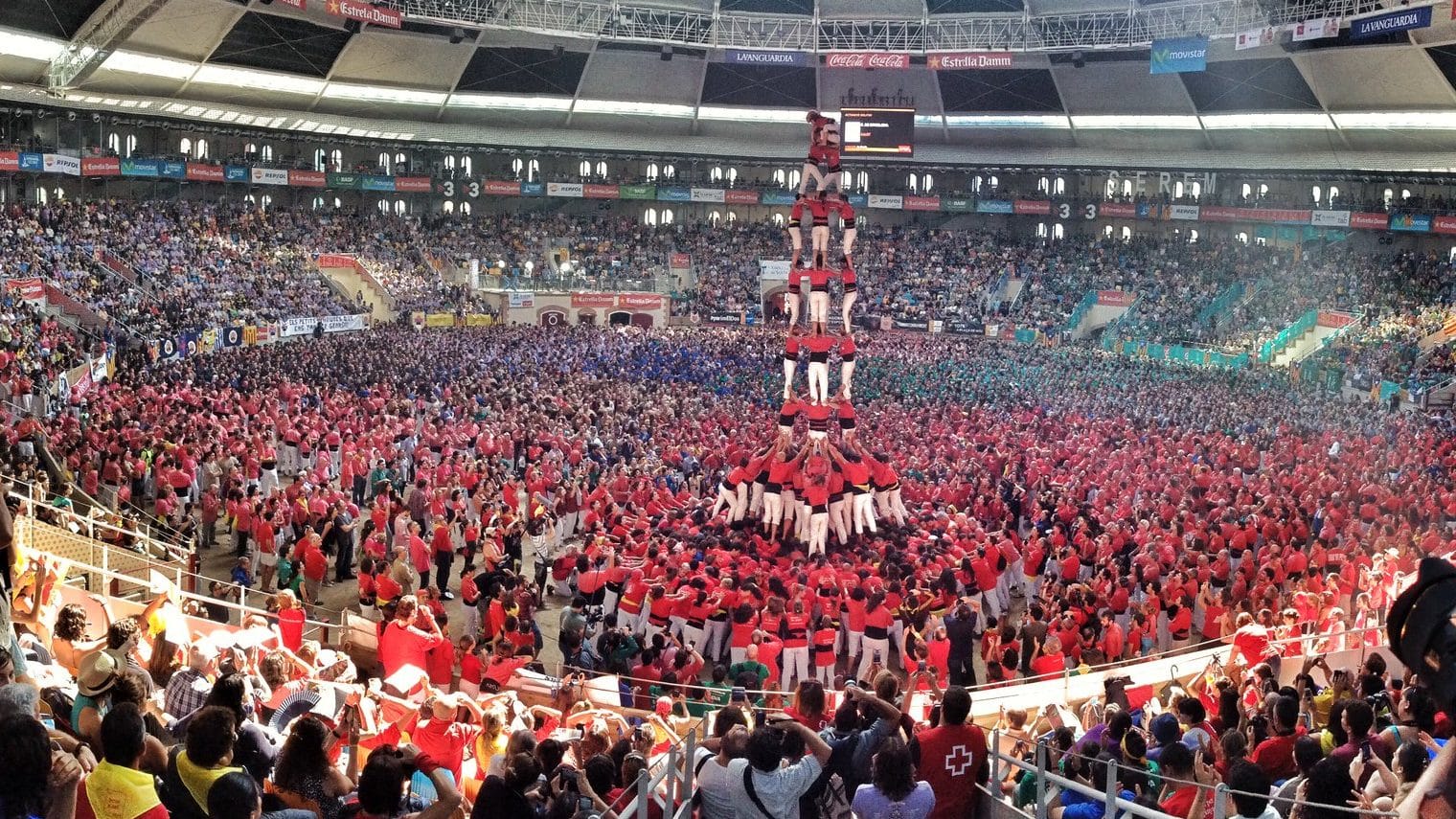 Image: a crowd of people watch as people build a human tower called a Castell