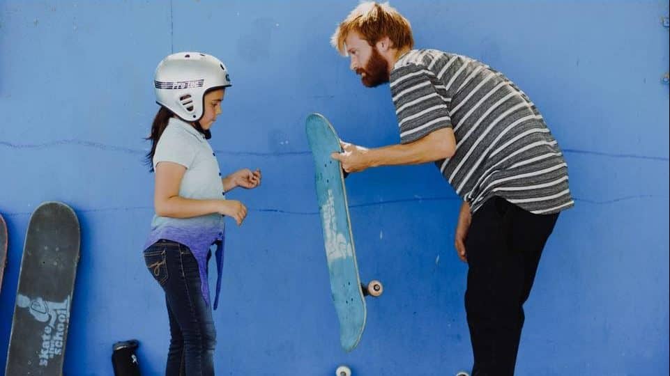 Image: Man handing a participant a skateboard that has the Skate After School logo on it