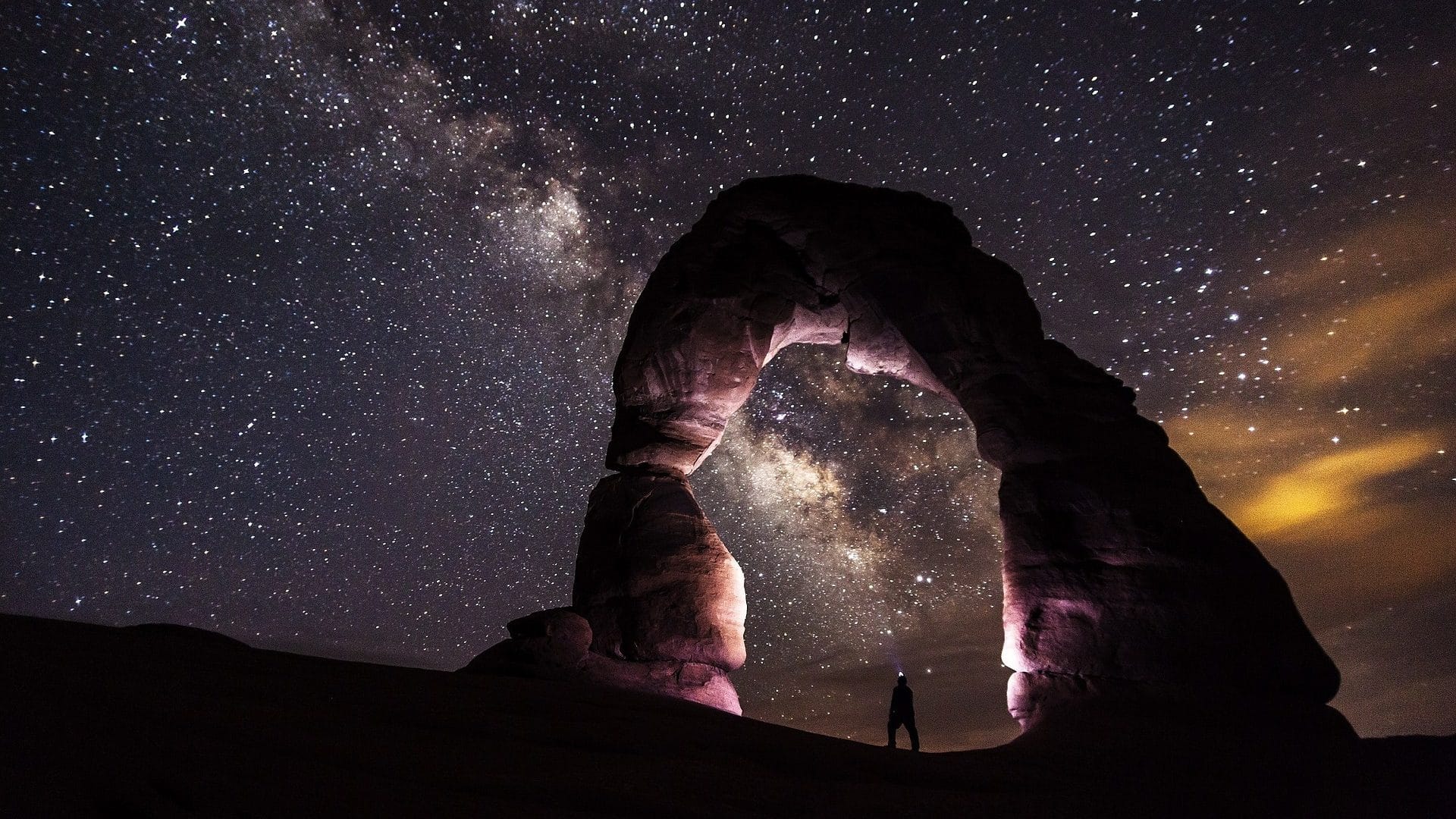 Image: Man standing underneath a rock arch looking up at the stars of the night sky with little to no light pollution.