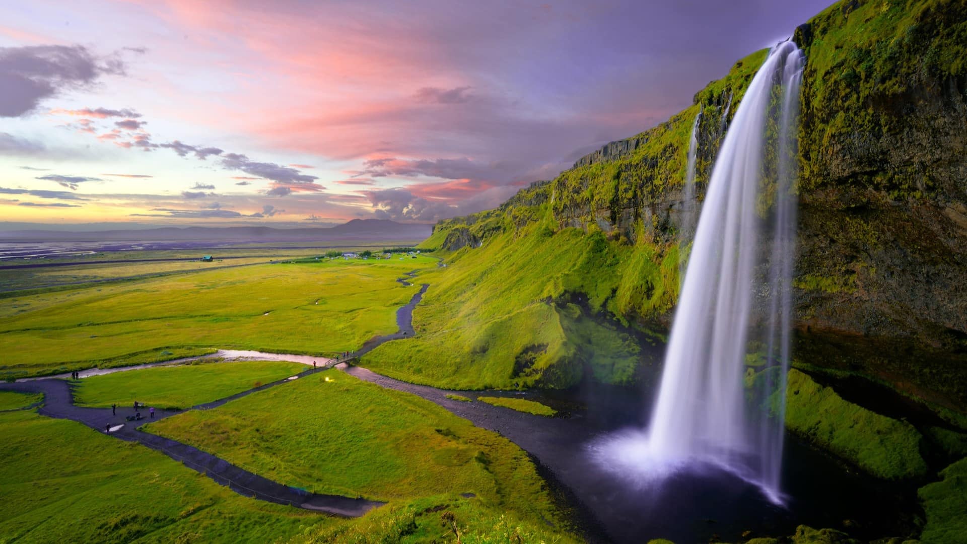 Image: A waterfall in Iceland sits in the foreground, with a striking sunset in the background. In the middle lies a vast area, once filled with lush forests, that is now a part of Mossy Earth's reforesting initiatives.