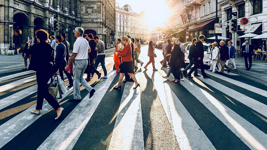 Image: a crowd of people walking across a street backlit by the setting sun
