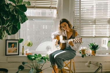 Image: A woman sitting by a window in a city apartment, playing guitar