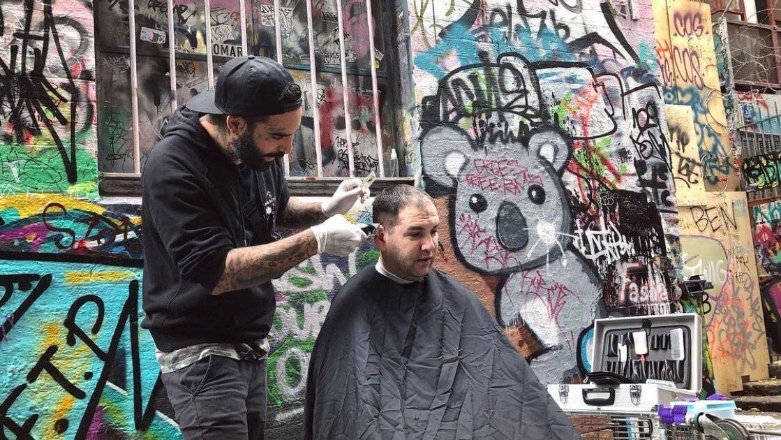 Image: The Streets Barber giving a haircut outside, next to a wall covered in beautiful graffiti