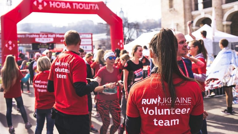 Image: A woman standing among volunteers at a charity foot race, facing away from the camera wearing a red shirt that says "volunteer team".