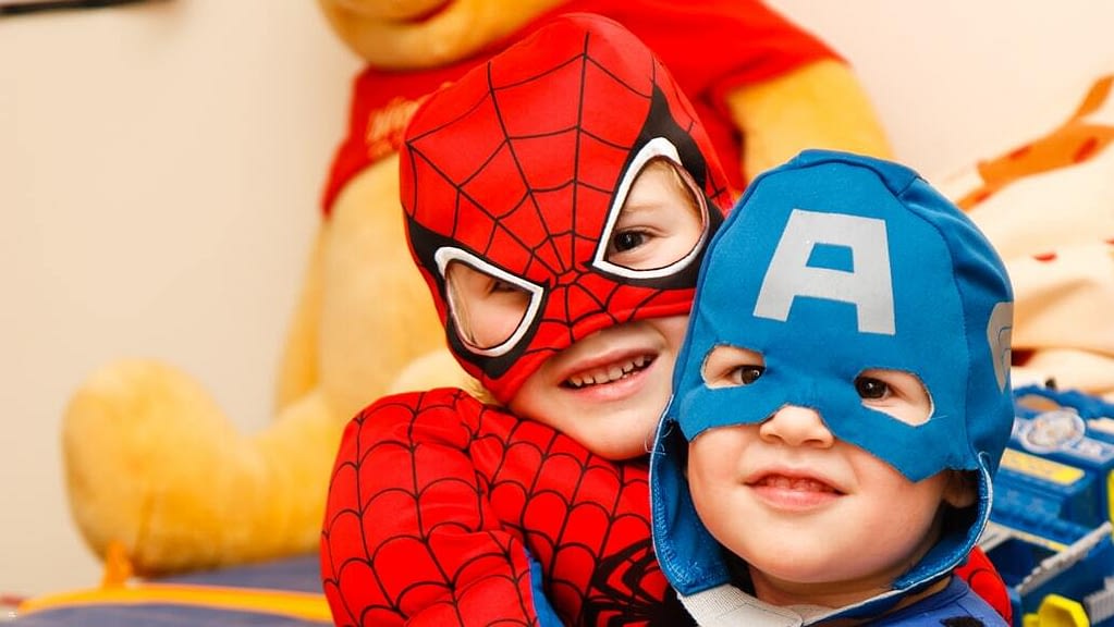 Image: Two kids smiling, one dressed as Spider Man and the other as Captain America. Kids with hearing loss are able to watch movies with this app.