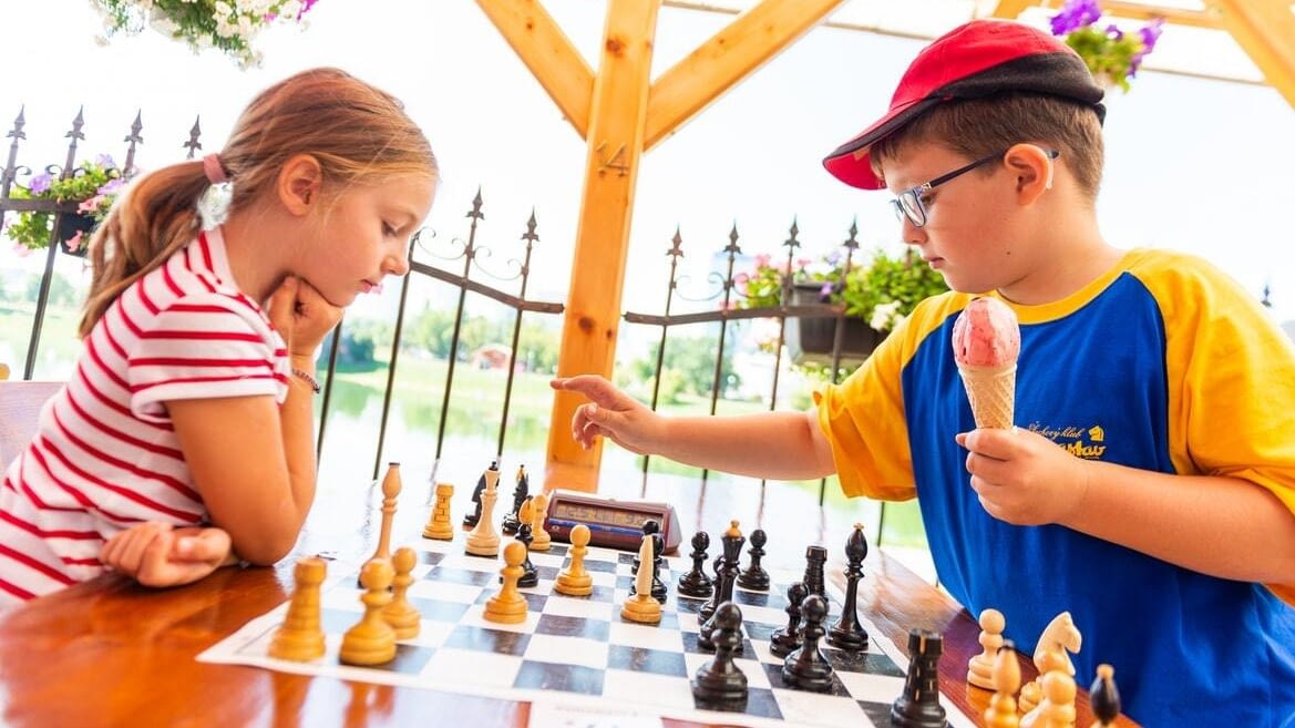 Image: two kids playing chess in a public park. On the left, a girl staring at the board, playing white. On the right, a boy holding an ice cream cone and making his move, playing black.