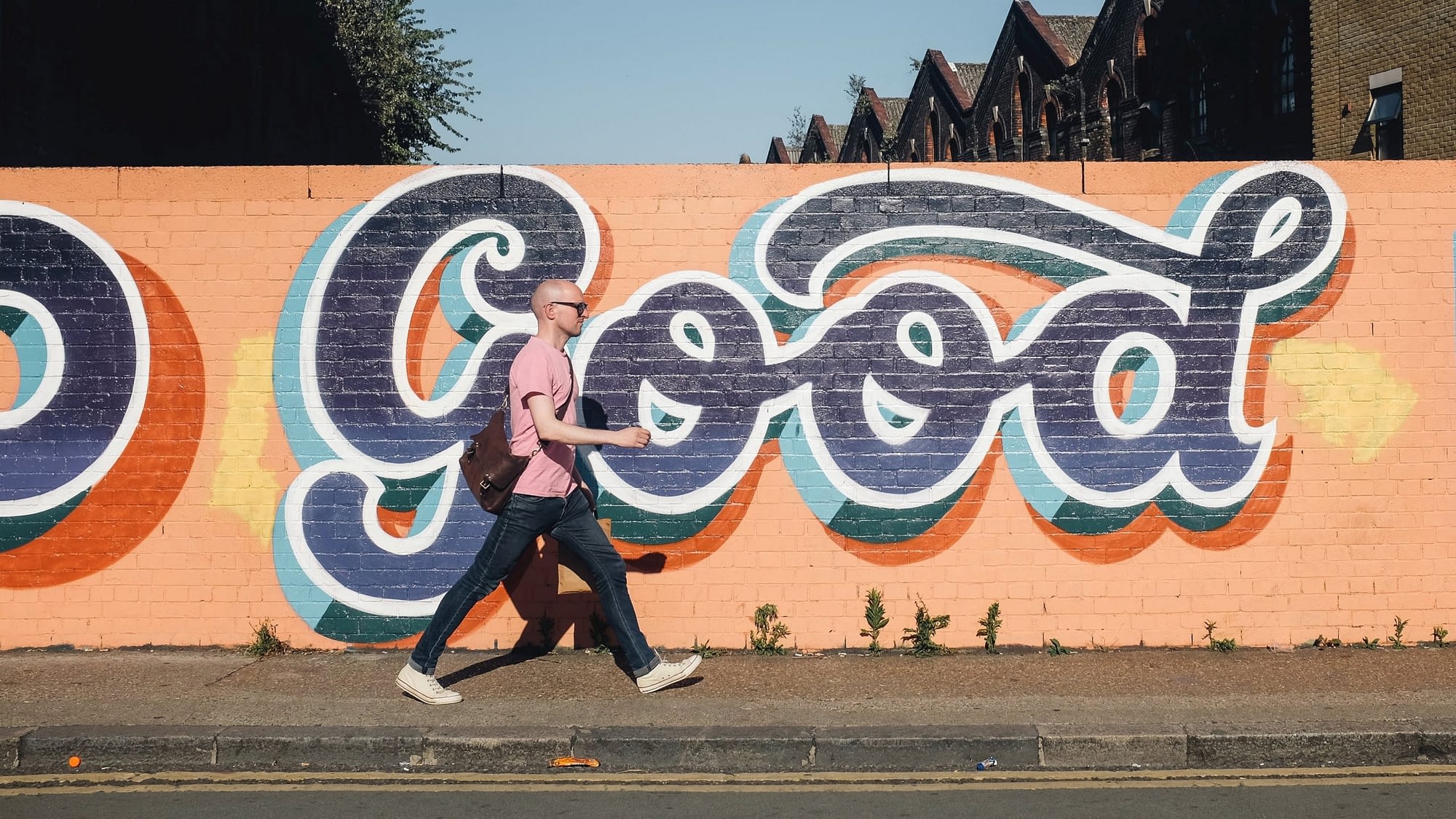 Image: A man walking down a side walk in front of an orange mural with blue writing that says "Good", using street wisdom practices.