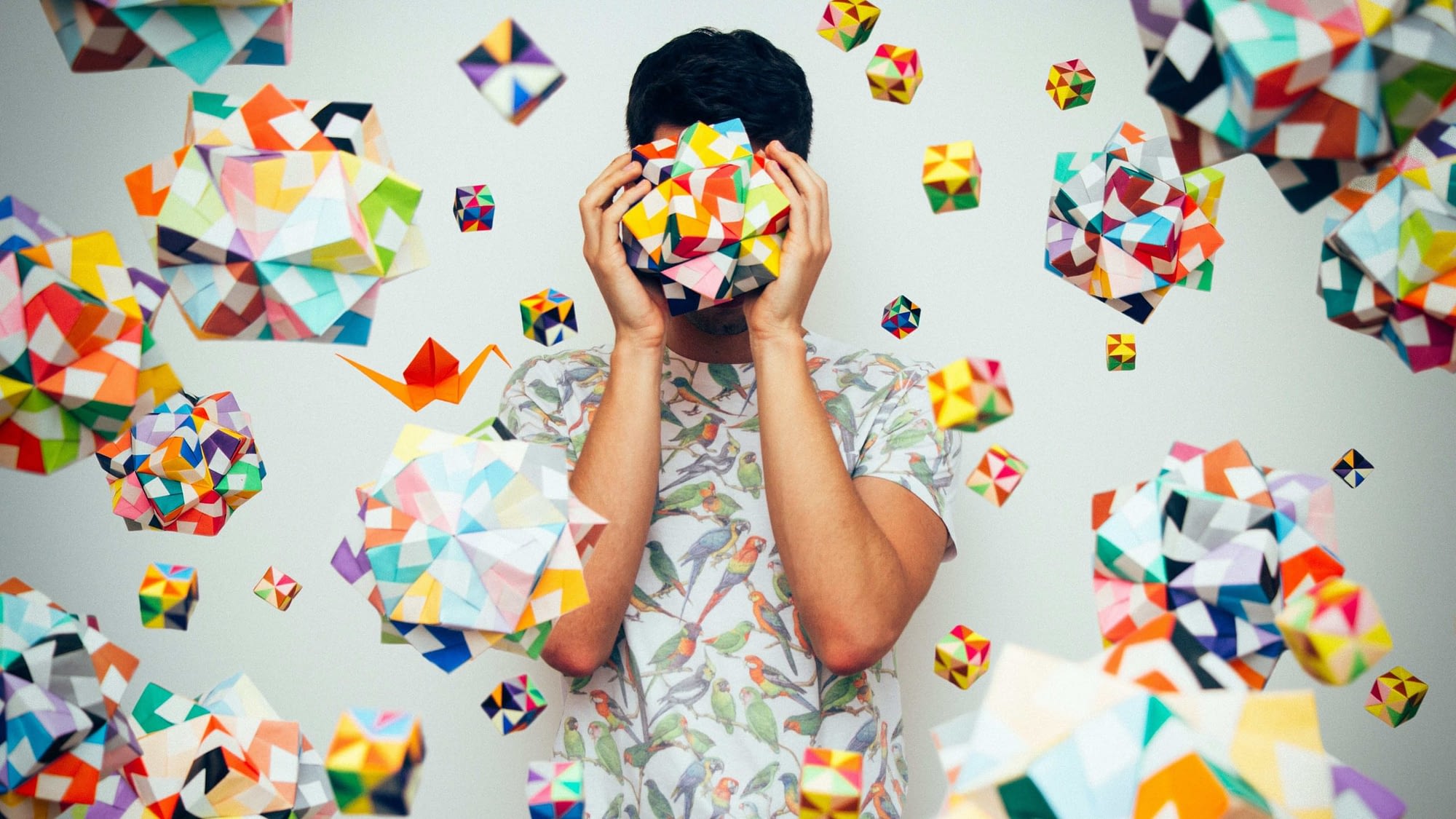 Image: A man holding a colorful cube in front of his face, and surrounded by hanging colorful cubes of the same type.