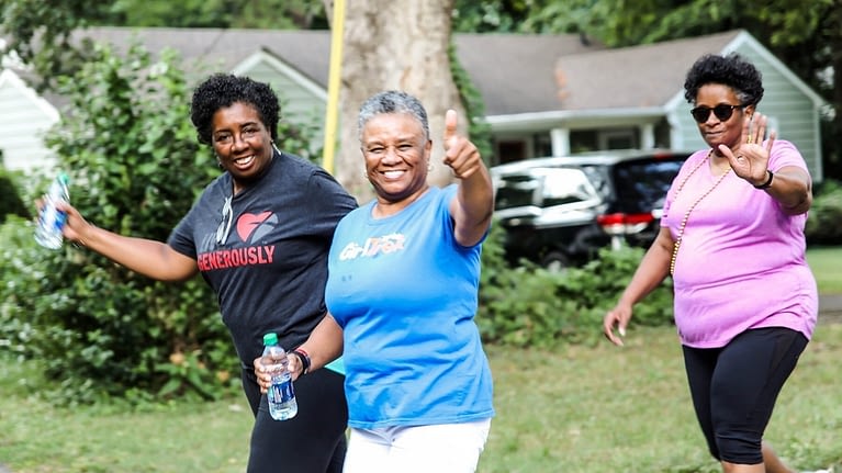 Source: Three older women smiling walking in a neighborhood with GirlTrek. One is giving a thumbs up, the others are waving