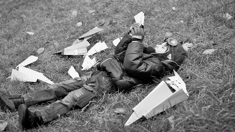 Image: Young child laying in the grass with paper airplanes all around him