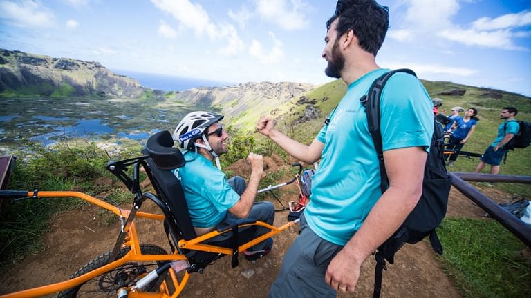 Image: Person in a trekking wheelchair fist bumping a friend atop a mountain