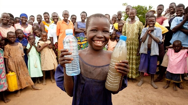 Image: child holds two water bottles one with clean healthy drinking water the other with brown muddy water, behind him are a crowd of people