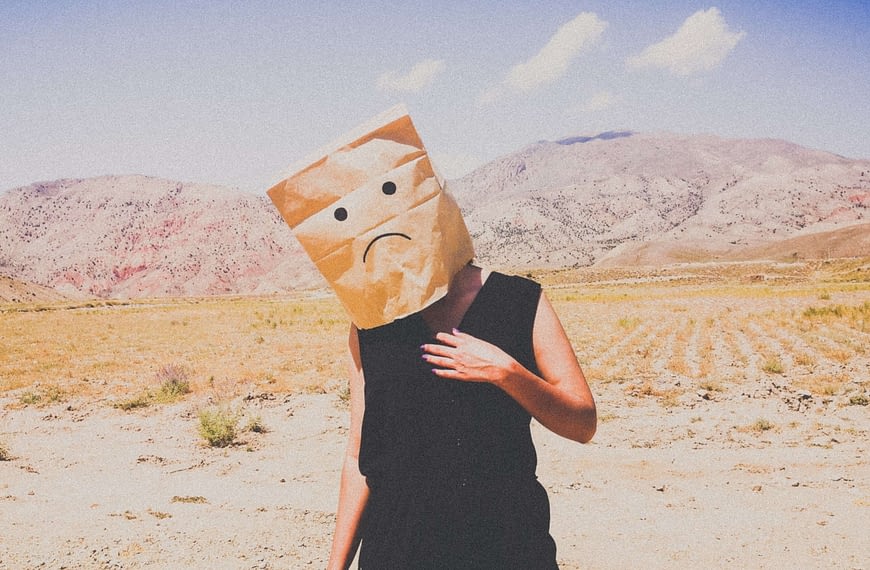 Image: A woman standing in a desert with a paper bag on her head, a frowning face drawn on the front. Symbolic of the emotions felt when we constantly say "I'll be happy when..."