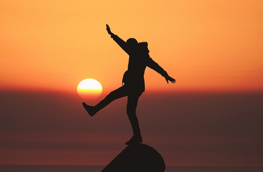Image: An individual standing on a peak with one leg and both arms extended in front of an orange sunset, symbolizing that there is no such thing as peace of mind.