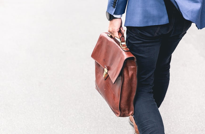 Image: A man walking away from the camera, holding a briefcase in his left hand with determination.