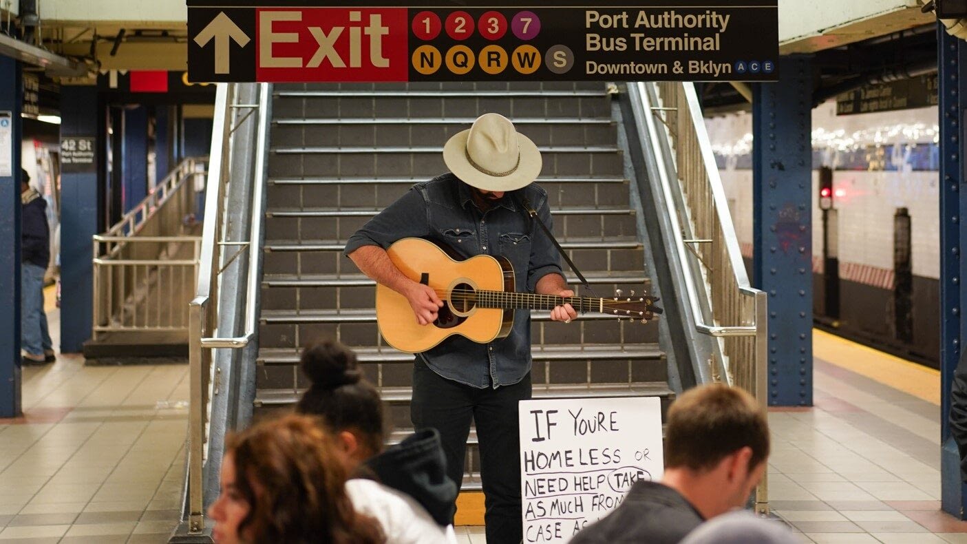 Image: Busker (Will) with Hopeful Cases playing guitar in the middle of a subway station with his head tilted down, next to the sign. 