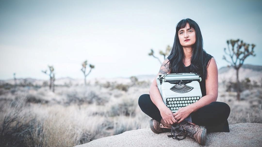 Image: Street Performer and poem writer Afrose Fatima Ahmed sitting on a rock in Joshua Tree holding her typewriter.
